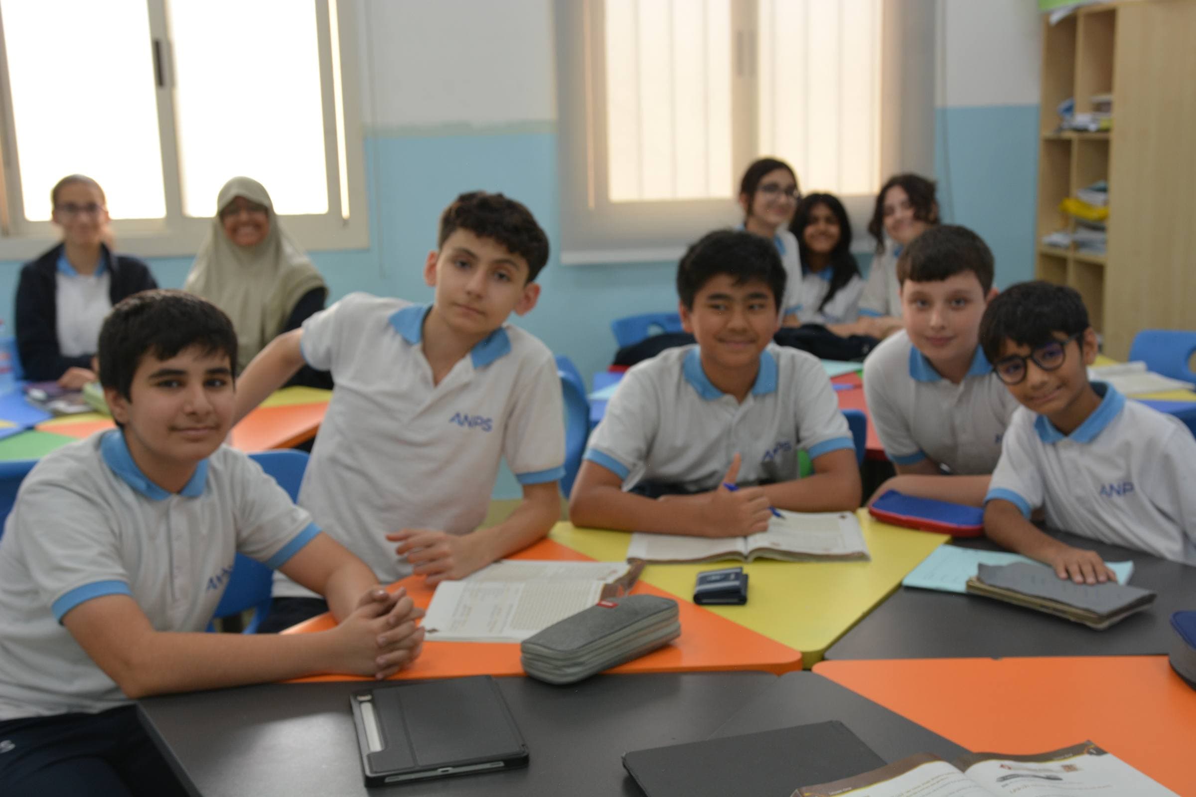 A bright classroom of Al Najah Private School students in the light-blue ANPS-branded uniform smiling together at their desks