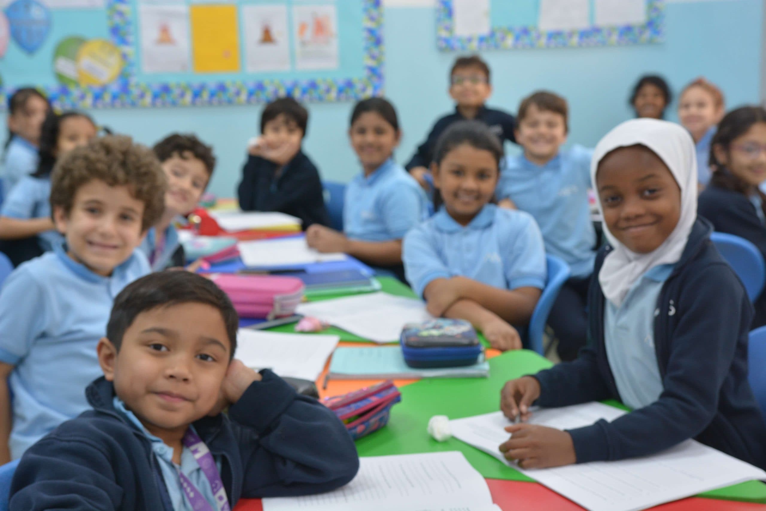 A diverse class of young Al Najah Private School pupils in the light-blue ANPS uniform smiling together at their colourful classroom desks